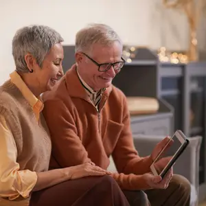 An elderly couple sits on a couch, the man wearing glasses and holding a tablet, and the woman wearing a beige sweater, both smiling and looking at the tablet