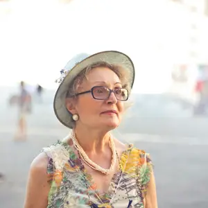 A woman wearing a hat, glasses, necklace, and earrings is standing on the street and looking to the left. The background is blurry and appears to be a street with some people walking in the distance. The light is coming from the left, illuminating the woman.