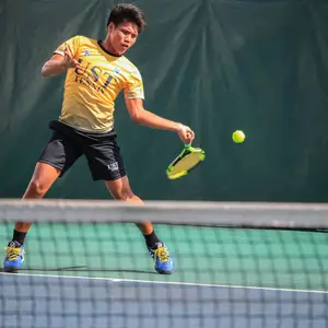 A man in a yellow shirt with UST written on it is playing tennis on a court.