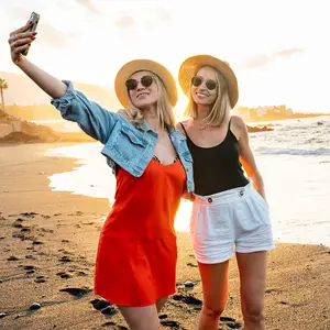 Two women are smiling on a beach and one is taking a selfie with her phone.
