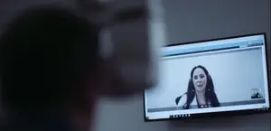 A woman wearing headphones is speaking into a camera on a monitor in a room with a blurry background