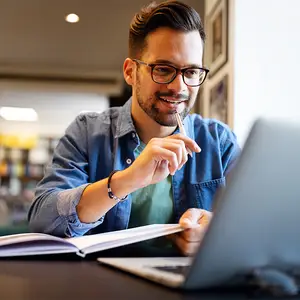 A smiling man is sitting at a desk in a library, holding a pen and a book, and using a laptop.