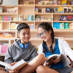Two kids are sitting in a library and reading books while smiling at each other