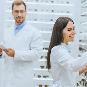A man and a woman in white lab coats stand in a room with white shelves holding glasses.