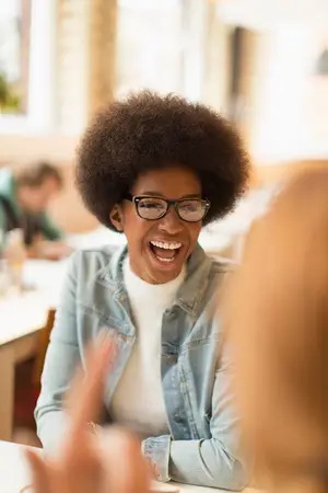 A woman with an afro hairstyle is laughing while wearing glasses and a denim jacket in a restaurant.