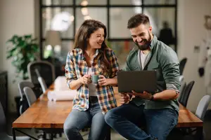 A man and woman sitting on a chair in an office, smiling and looking at a laptop
