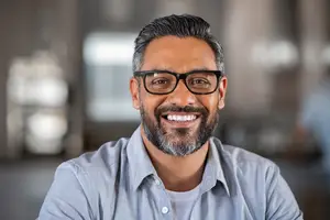 Portrait of a smiling man in glasses and a blue shirt looking at the camera in an office environment