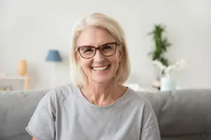 A woman with short blonde hair wearing glasses is sitting on a couch and smiling at the camera