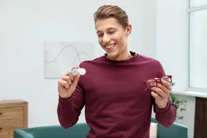 A man holds a contact lens in his left hand and a pair of red-framed glasses in his right hand, while smiling and standing in a room with a white wall, a wooden cabinet, and a green couch.