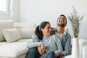 A man and woman sitting on a white couch, laughing and looking at the camera.