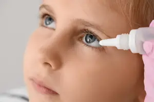 A young girl with light pink gloves administering eye drops into her eye