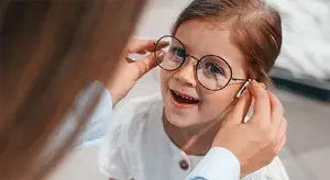 A young girl with glasses and a smiling face is having her glasses adjusted by a woman wearing a white shirt