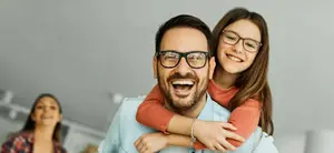 A man with a girl sitting on his shoulder, they are both smiling, and a woman is standing behind them in a room with white walls and a ceiling.