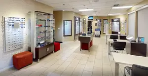 An optical store with a variety of eyewear on display, including glasses and sunglasses, arranged on shelves and tables, with a comfortable seating area, and digital signage displaying promotional offers. The space is well-lit with ceiling-mounted lamps and features a clean, modern design with beige walls and tiled flooring. The store also includes a monitor on the wall for displaying information or advertisements.