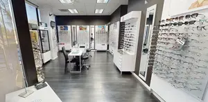 An optical shop displays a variety of glasses on a wall, with a desk, chairs, and a phone. The shop has a modern design with a black wall and white shelves. The glasses are arranged in rows, and some are on a table. The shop has a bright and airy atmosphere, with a large glass window on the left side. The shop is empty, and there are no people in it.
