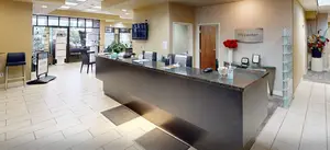 Image of an eye care center lobby with a front desk, a television, and a vase with flowers on a table, taken during the daytime.