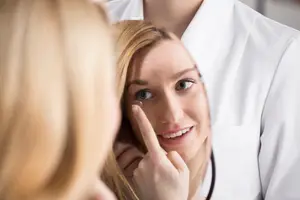 A woman is looking at her eye in a mirror while wearing a contact lens and a man in a white coat is looking at her from the side