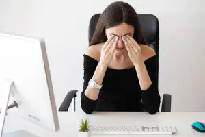 A woman sitting at a desk with a computer monitor covering her eyes with her hands.