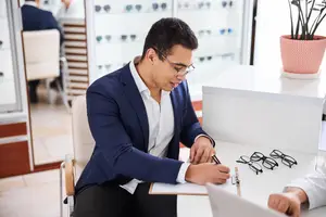A man in a suit and glasses is writing on a clipboard at an optometrist's office.