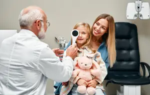 A doctor is examining a young girl's eye with a flashlight while a woman and a stuffed animal watch nearby.