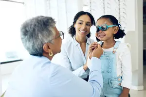 A young girl wearing eye glasses is being checked by a doctor with a mother watching in the background