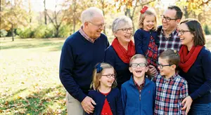 A family of seven people are standing in a park for a family photo, smiling for the camera. They are all wearing sweaters and scarves, with the two adults on either side of the group. The background is blurry with trees and grass.