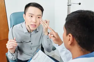Asian male patient consulting with doctor about eye condition, possibly dry eye syndrome, in a medical office setting.