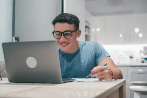 A man is smiling and seems to be working on his laptop while sitting on a chair in front of a table in the kitchen.