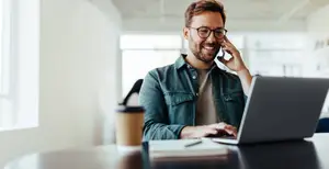 A smiling man wearing glasses is talking on his cellphone while working on his laptop at his desk in an office room.