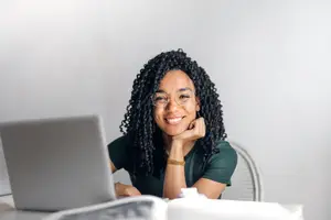 A smiling woman with curly hair working on a laptop in a room with white walls