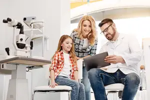 A young girl is sitting on a chair, smiling and looking at a man and a woman who are holding a tablet and looking at it. The man is wearing glasses and a white lab coat. Behind them is a white machine with a black handle, probably a microscope, and a glass bottle on a table.
