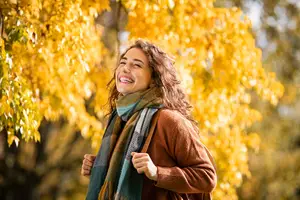 A smiling woman with curly hair wearing a brown sweater and a scarf stands under a tree with yellow leaves on a sunny day