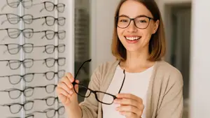 A woman with short hair is smiling and holding glasses in front of her face in a room with many glasses displayed on a white wall.