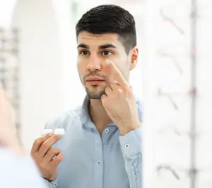 Man inserting a contact lens into his eye in front of a mirror.