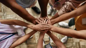 A group of people from different backgrounds are placing their hands on top of each other in a circle on a tiled floor, wearing sneakers and ID cards, forming a symbol of unity and collaboration.
