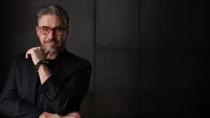A man with glasses and a black suit jacket is posing for a picture in front of a dark background