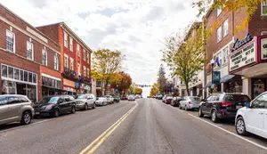 A city street with multiple cars parked on either side, with a clear sky and some clouds in the background.