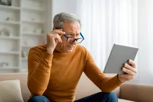 An older man in an orange sweater is holding a tablet and looking at it while sitting on a couch.