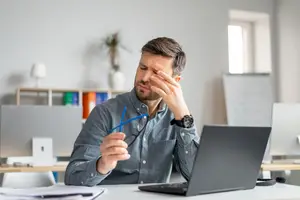 A man sitting at a desk holding glasses and rubbing his eyes