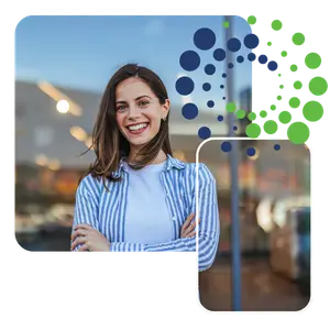 A smiling woman with brown hair is standing in a shop, wearing a blue and white striped shirt and a white shirt underneath, posing for a photo with her arms crossed in front of her chest.