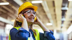 A smiling woman wearing a yellow hard hat, safety glasses, and headphones is standing inside a factory building, likely a supervisor or safety officer.