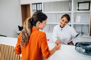 Two women at a clinic's reception area, one wearing an orange shirt and the other a white lab coat, smiling at each other.