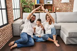 A family of three, with a young boy, a father, and a mother, are sitting on the floor and smiling while raising their hands in the living room