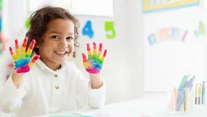Little girl showing her rainbow-colored painted hands at a table in a classroom.