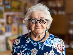 An elderly woman with white hair and glasses smiles at the camera while standing in front of a wall with various paintings.