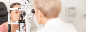 A young woman with brown eyes having her eyes examined by an optometrist in a white coat using a slit lamp.