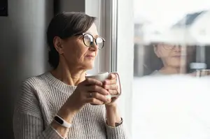An elderly woman with glasses and a sweater drinks coffee while looking out of a window with a reflection of another woman