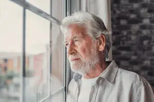 An old man with white hair and beard is looking out of the window with a serious expression