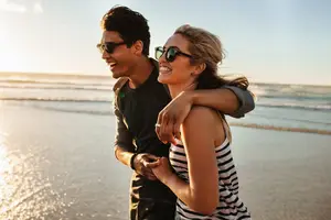 A man and woman smiling on the beach during sunset with the ocean in the background.