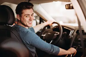 A smiling man in a blue shirt is driving a car with his right hand on the steering wheel.